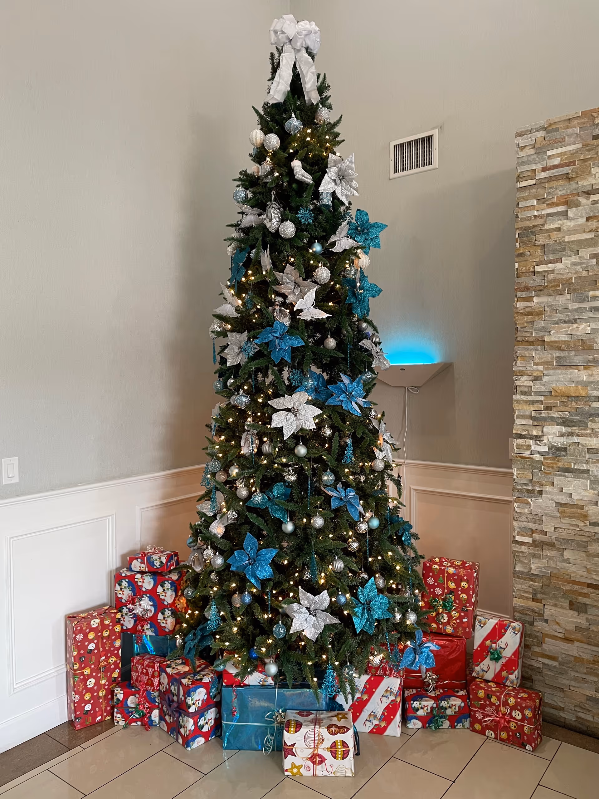 Tall decorated Christmas tree with blue and silver ornaments and wrapped presents at its base in an interior lobby.