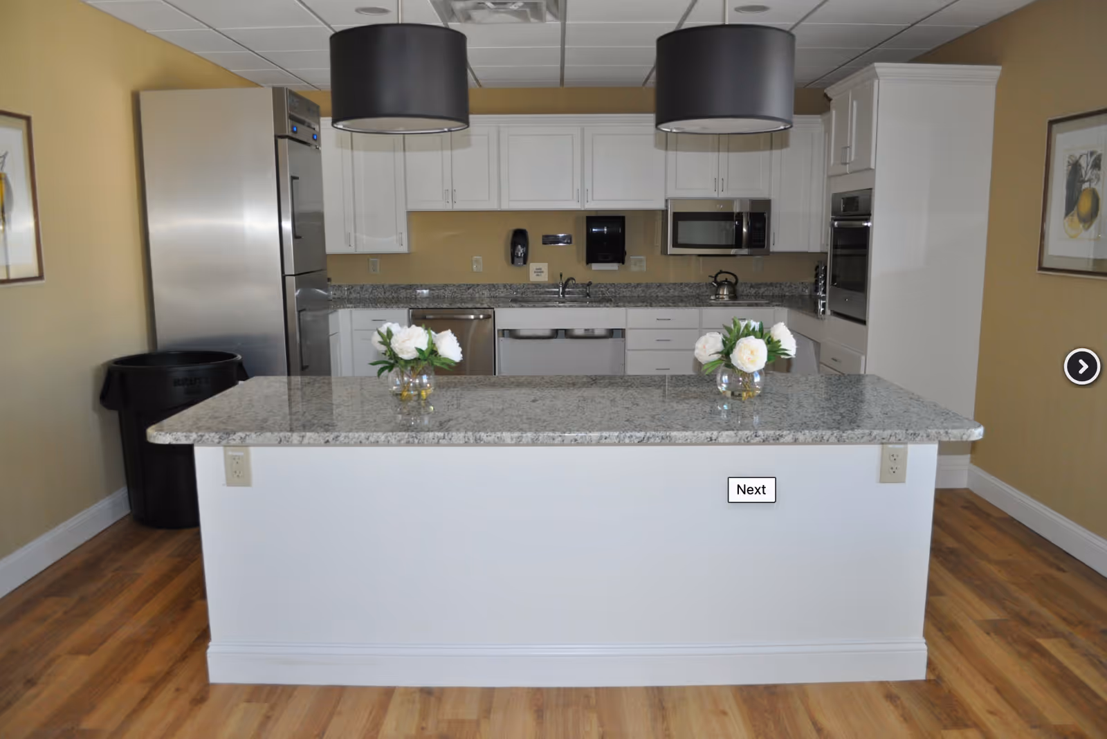 Modern communal kitchen with a granite island, stainless steel appliances, pendant lights, and two small vases of flowers.