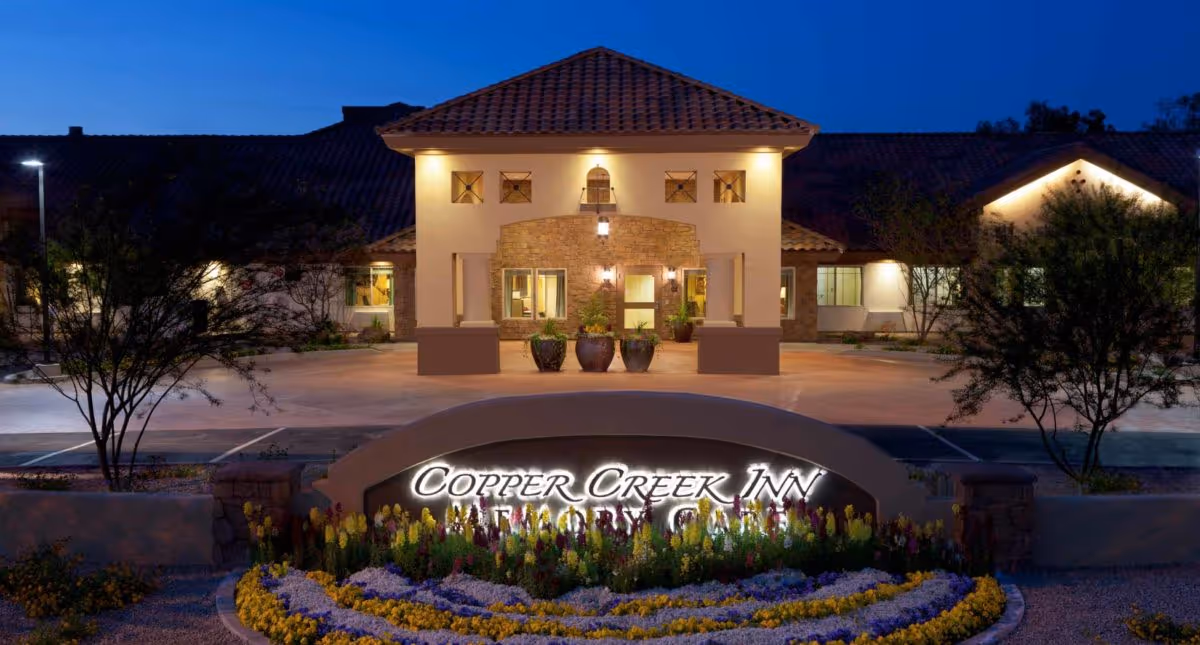 Night view of the entrance to Copper Creek Inn Memory Care facility with illuminated sign, landscaped flower beds, and a building with warm exterior lighting and a tiled roof.