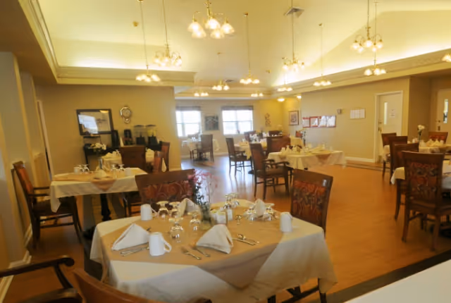 A well-lit dining room with multiple tables set for a meal, featuring white tablecloths, folded napkins, glassware, and silverware. The room has wooden chairs with patterned upholstery, warm beige walls, and several ceiling light fixtures. Windows in the background allow natural light to enter the space.