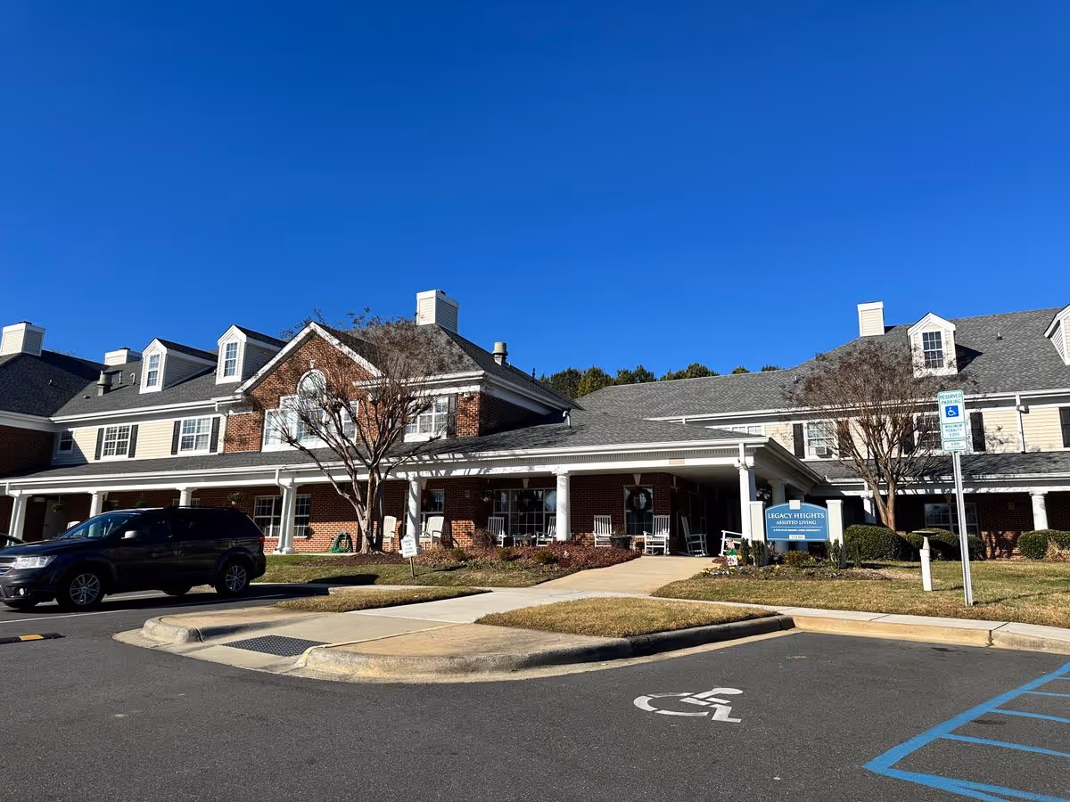 Front exterior of Legacy Heights Senior Living Community building with a covered entrance, rockers on the porch, parked car, and a visible accessible parking space under a clear blue sky.