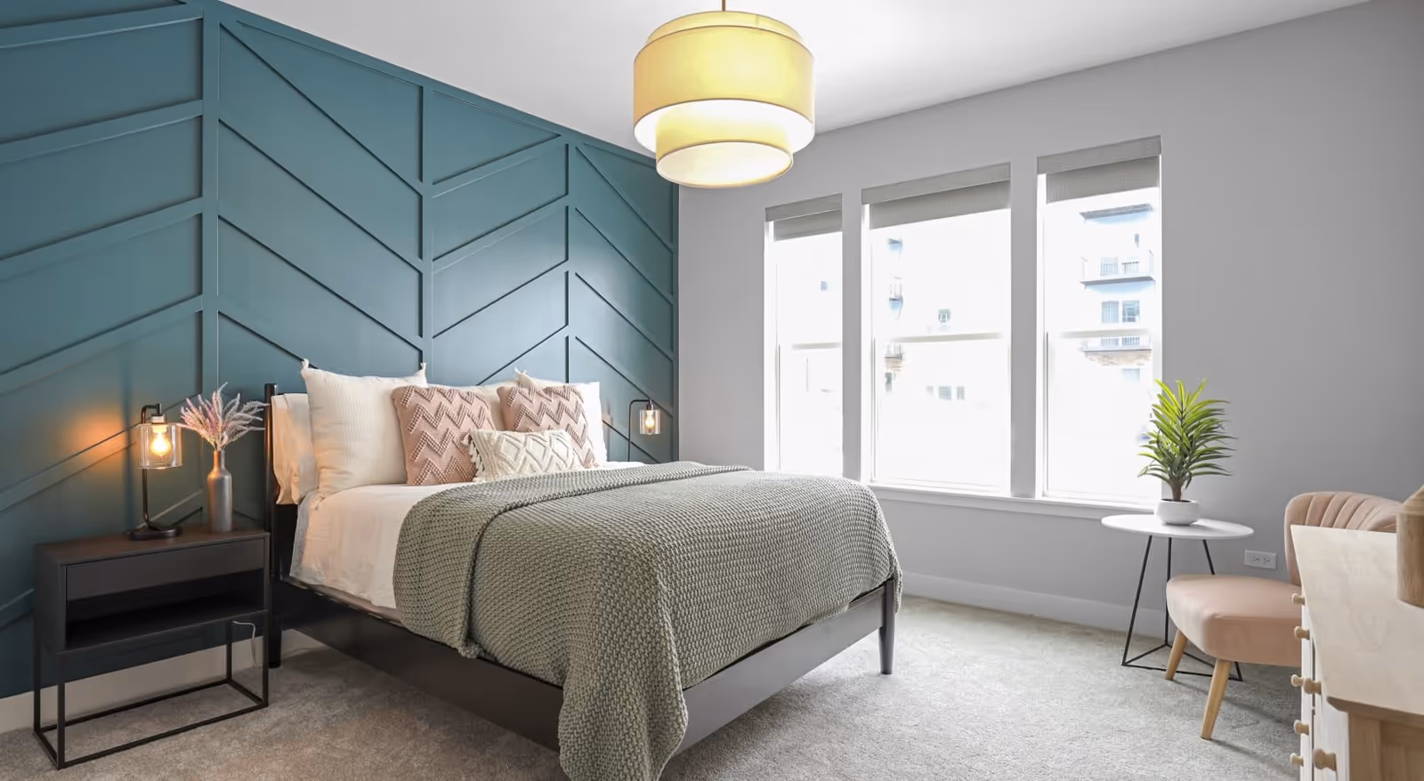 A modern bedroom with a dark wood bed frame, a green textured blanket, and several decorative pillows. The wall behind the bed features a geometric pattern painted in teal. There is a black nightstand with a lamp and a vase with flowers on the left side of the bed. Three large windows with blinds allow natural light to fill the room. On the right side, there is a small round white table with a potted plant and a light pink upholstered chair. A light wood dresser is partially visible on the right.