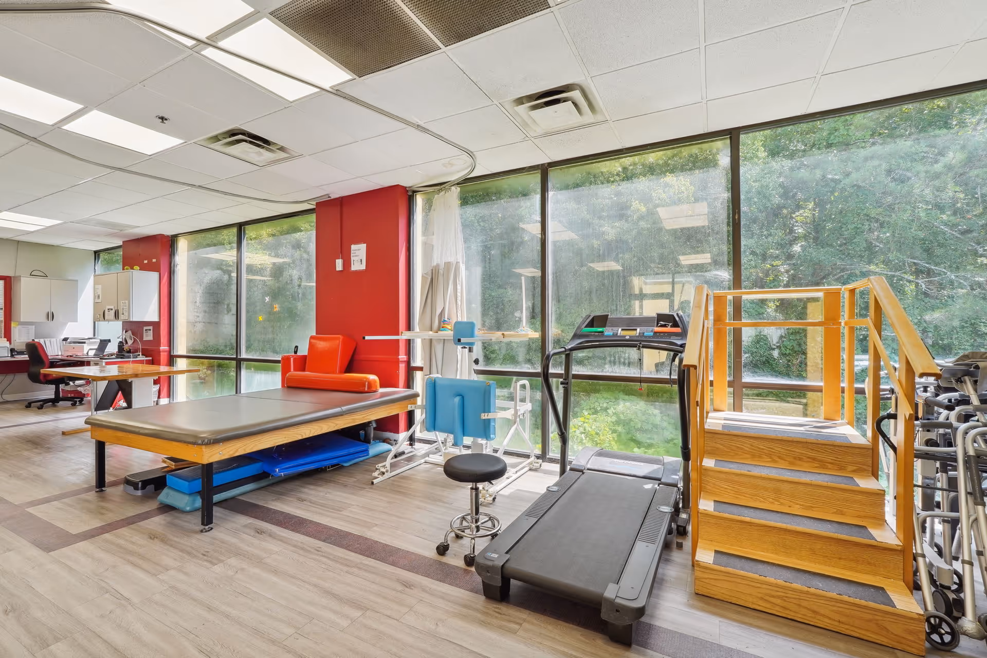 A bright physical therapy room with large windows overlooking greenery. The room contains a treadmill, a set of wooden stairs with handrails for rehabilitation, a padded therapy table, a red armchair, and various therapy equipment. The floor is wood-patterned, and the ceiling has fluorescent lights.