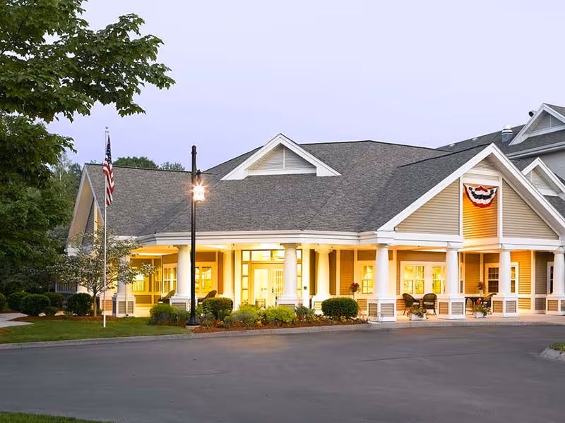 Front entrance of a senior living facility with a covered porch, white columns, warm interior lighting, an American flag, and landscaped beds.