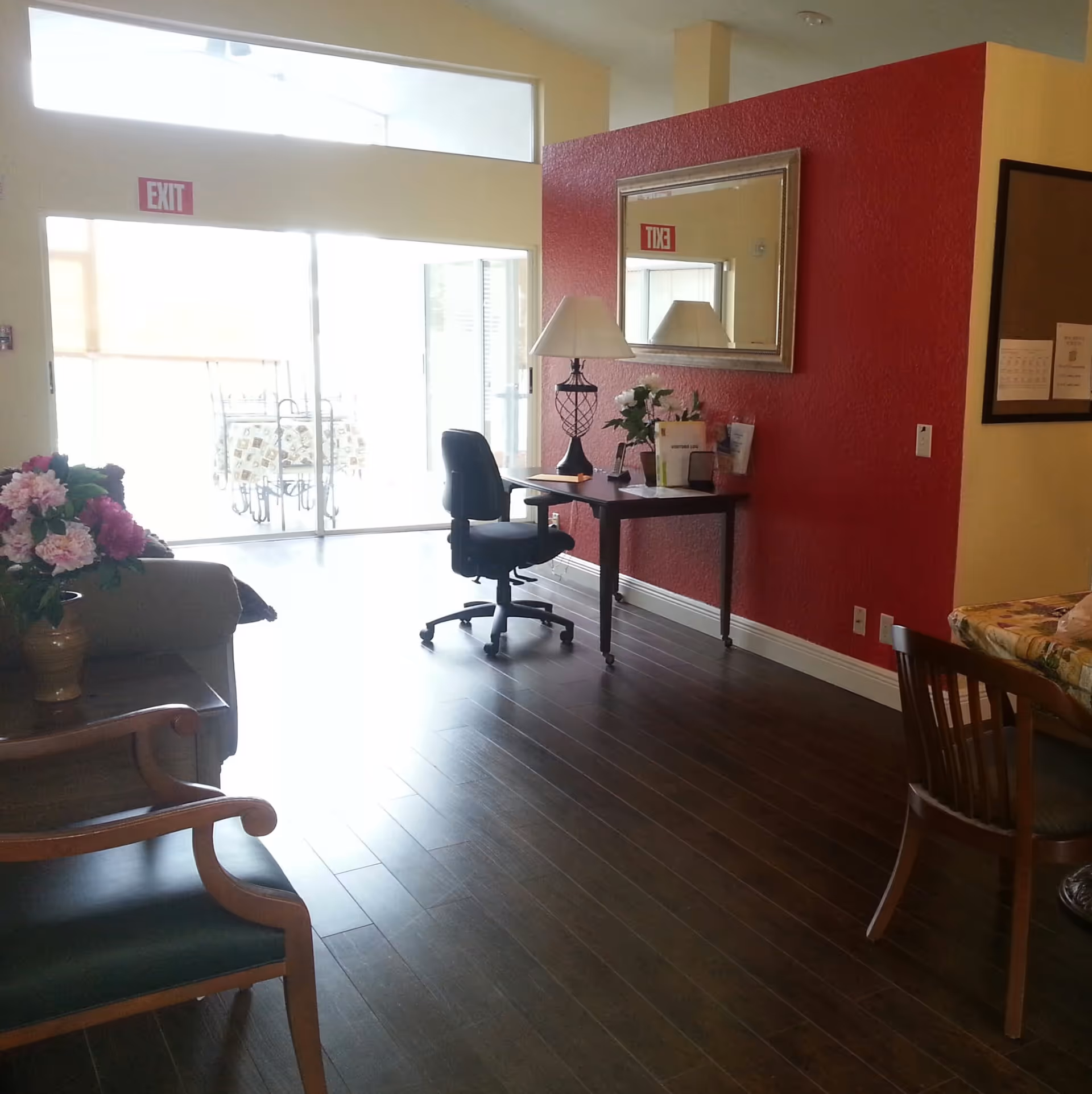 Well-lit senior living common area with hardwood floors, a desk and office chair against a red accent wall, seating and a floral arrangement near sliding glass doors.