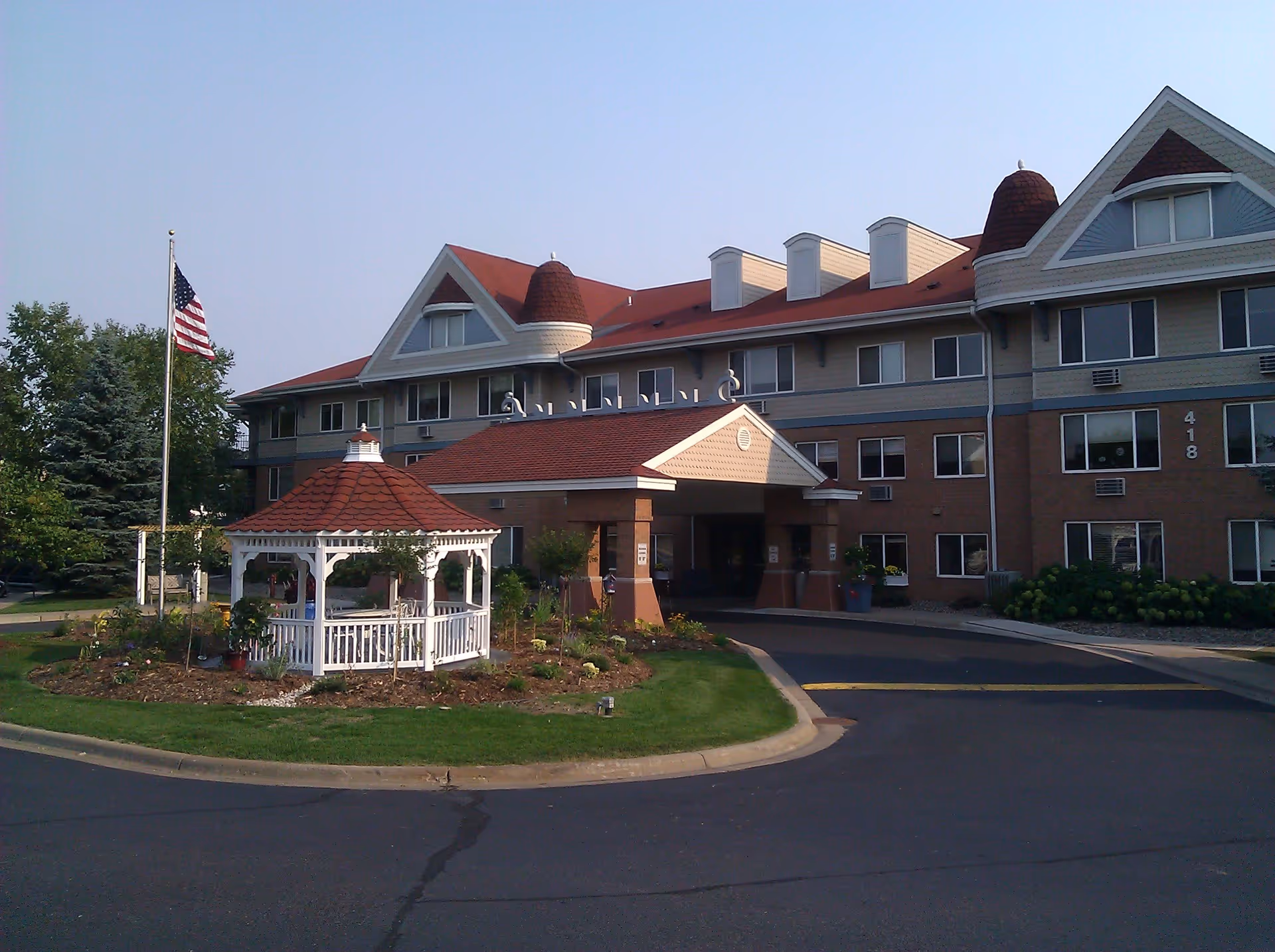 Exterior view of a multi-story senior living facility with a red roof and beige siding. In front of the building is a white gazebo surrounded by a landscaped garden and an American flag on a flagpole. The driveway curves around the garden area leading to the building entrance.