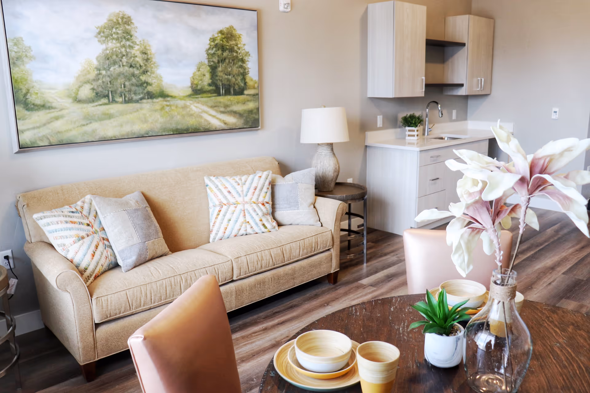 Bright living area with a beige sofa and patterned pillows, wall painting, small kitchenette, and a dining table with a vase of flowers.