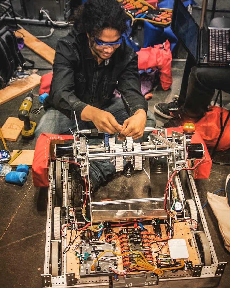 Person wearing safety glasses assembles a metal robotic frame in a cluttered workshop.