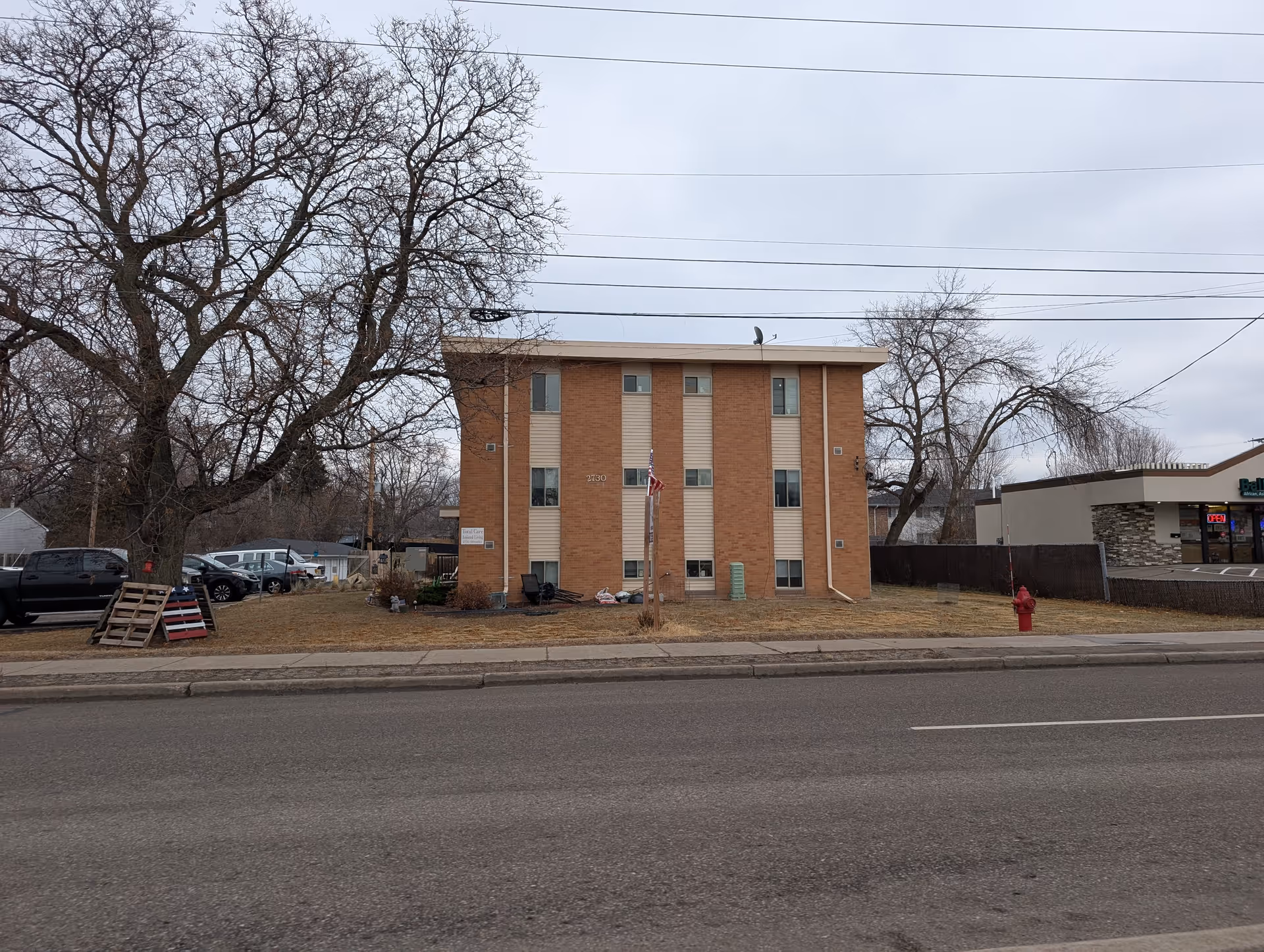Three-story brick apartment building with a flagpole on its front lawn, flanked by leafless trees and a street in the foreground.