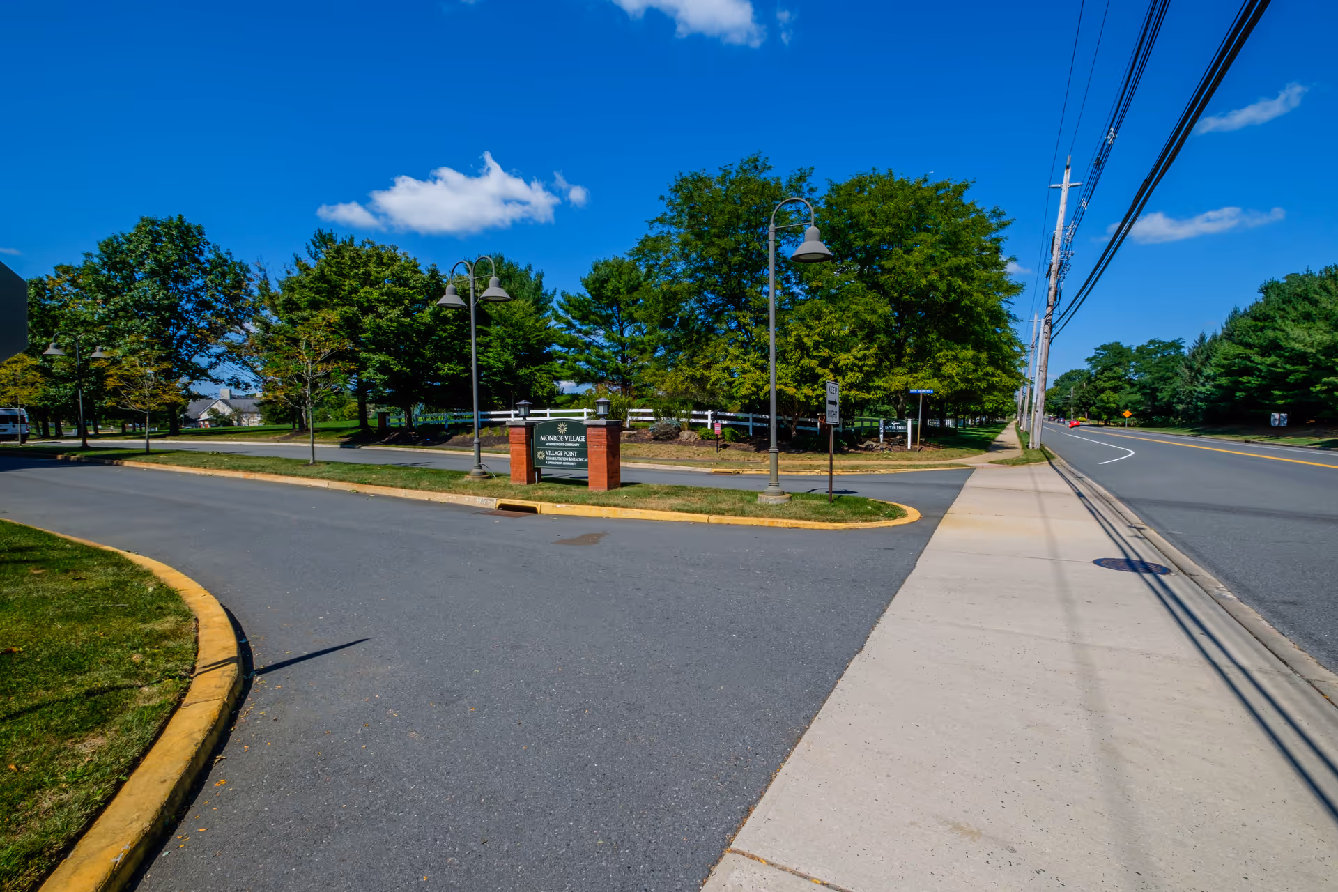 Entrance drive and sign for Monroe Village with lamp posts, trees, a sidewalk and road under a blue sky.