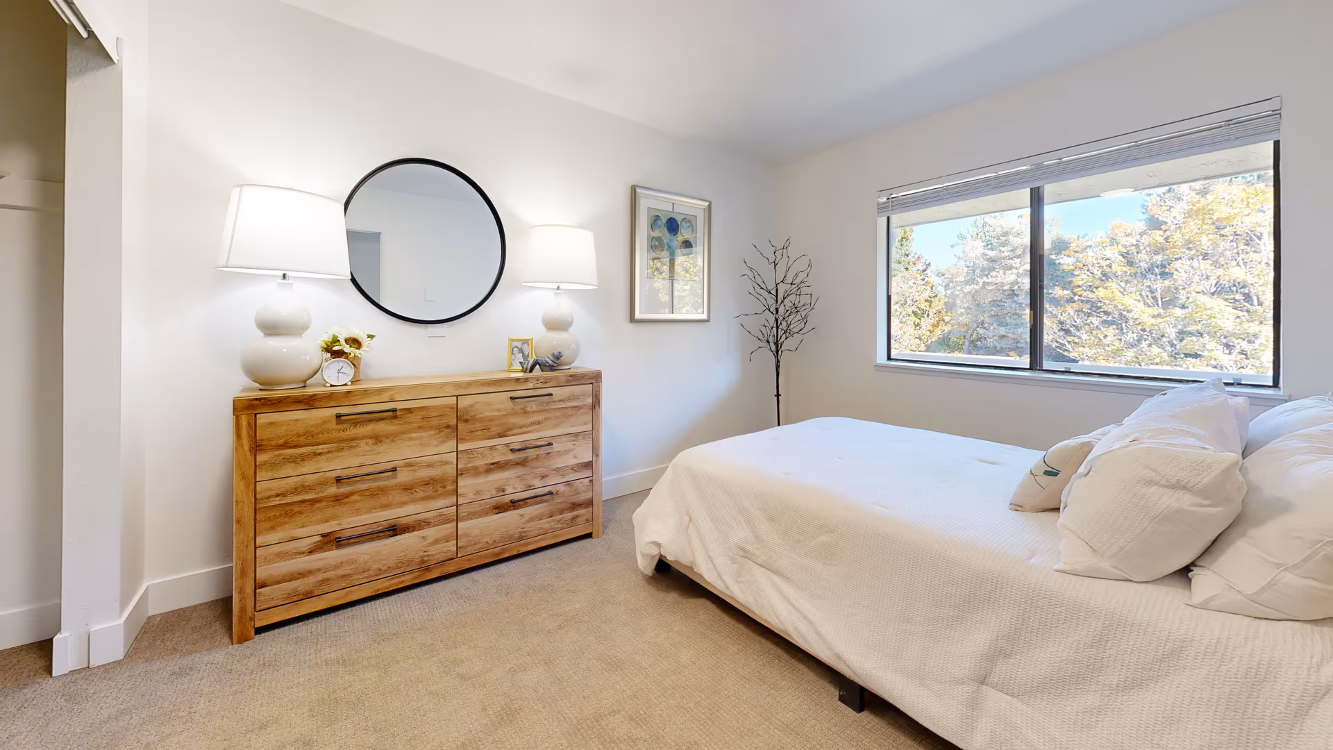 Bright bedroom with a wooden dresser, round mirror, lamps, and a large window.