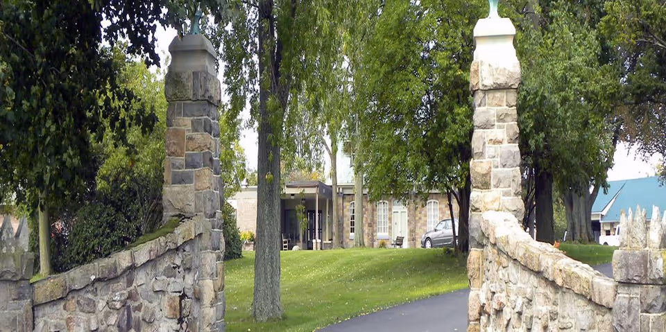 Stone entrance pillars with a curved stone wall on each side leading to a paved driveway surrounded by green grass and trees, with a building partially visible in the background.