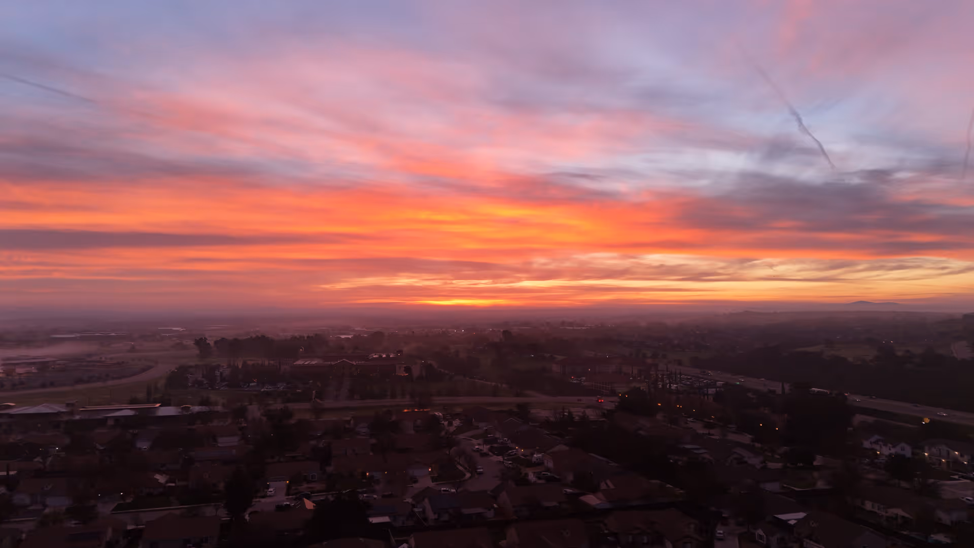 Colorful sunrise over a suburban neighborhood and distant landscape seen from above.
