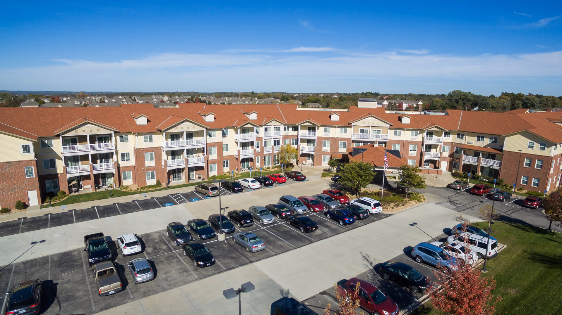 Aerial view of a large senior living facility named Shawnee Hills with a three-story building featuring balconies and a red roof. The parking lot in front is partially filled with cars, and there are trees and landscaped areas around the building under a clear blue sky.