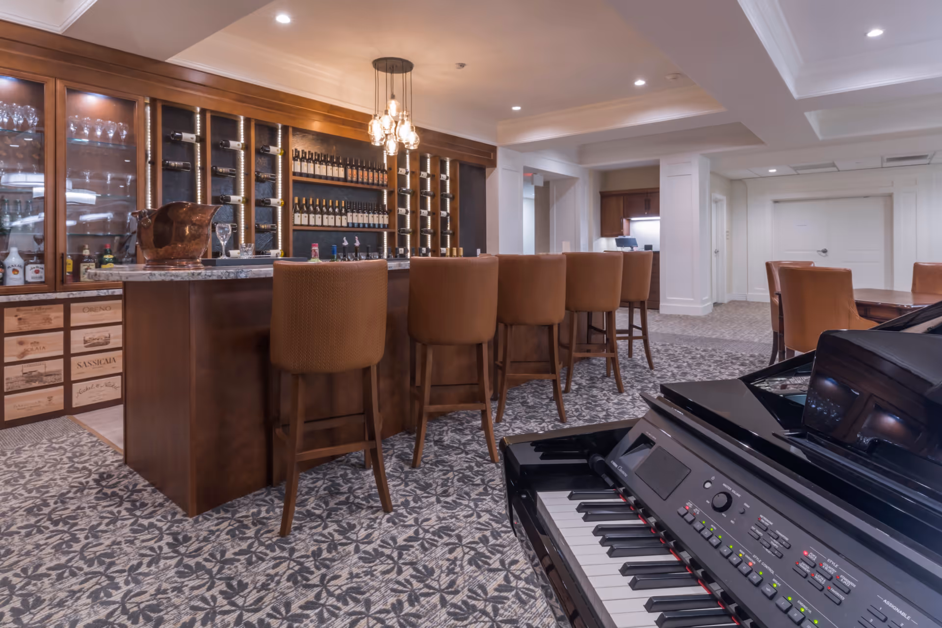 Interior view of a senior living facility lounge area featuring a bar with several brown cushioned bar stools, a wine rack with bottles, glassware displayed in cabinets, and a black digital piano in the foreground. The room has patterned carpet flooring and recessed lighting in the ceiling.