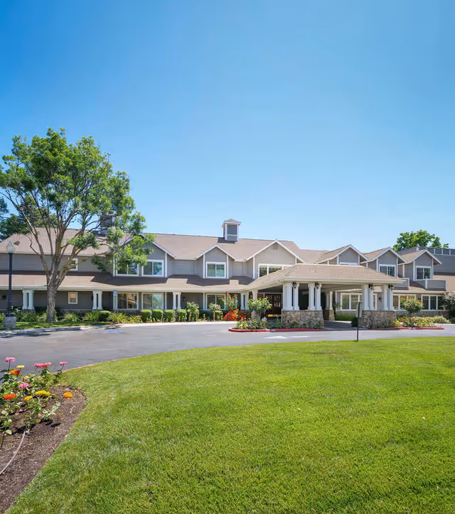 Front exterior view of The Grove Senior Living facility with a large green lawn, flower beds, a tree, and a covered entrance under a clear blue sky.