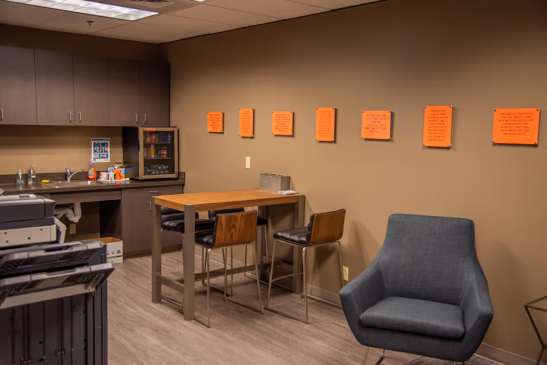 Cozy staff lounge with kitchenette, high table, stools, gray chair, and orange notes on beige wall.