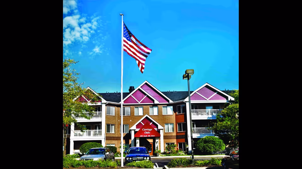A multi-story Carriage Oaks senior living building with purple accents and an American flag.