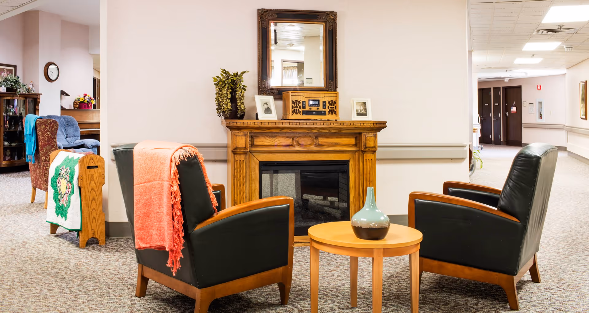 A cozy seating area in a senior living facility hallway with two black leather armchairs with wooden frames, each draped with a blanket. Between the chairs is a small round wooden table with a decorative vase. Behind the chairs is a wooden electric fireplace mantel with a mirror above it, flanked by framed photos and a vintage-style radio. The hallway is carpeted and decorated with plants and furniture, with doors visible in the background.