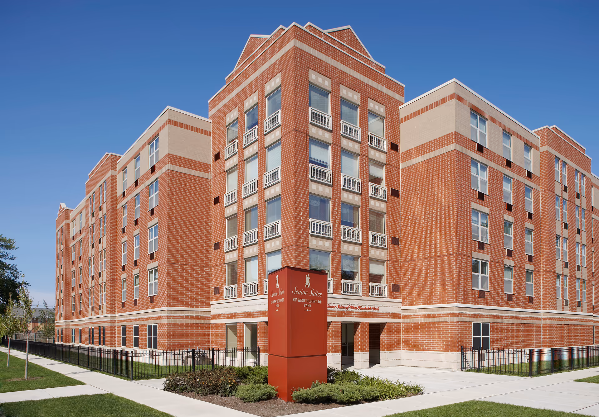 Exterior view of a multi-story brick building with large windows and a red sign in front that reads 'Senior Suites of West Humboldt Park' under a clear blue sky.