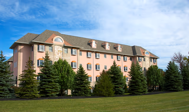Pink three-story senior living building with tall evergreen trees against a blue sky.
