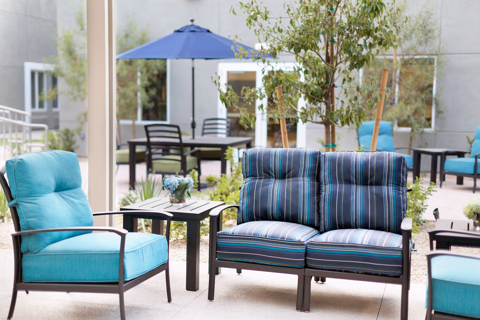 Outdoor patio area with cushioned seating including a turquoise armchair and a striped loveseat, a small table with a flower arrangement, additional tables and chairs in the background, a blue umbrella, and some greenery and trees around.