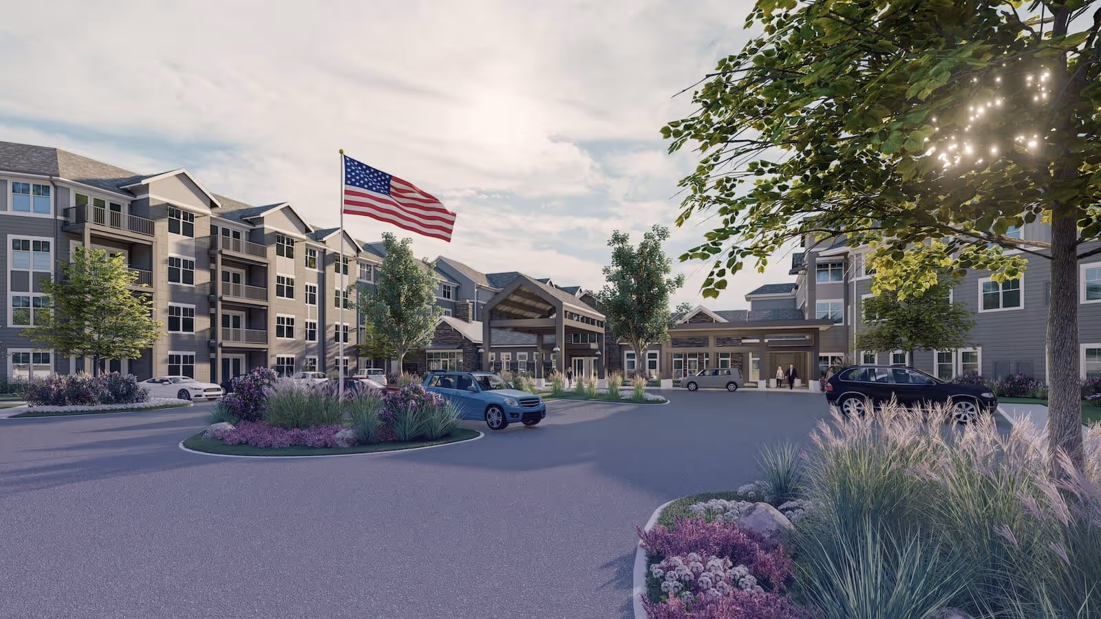 Front entrance of a multi-story senior living building with a circular drive, American flag, landscaped beds, and parked cars.