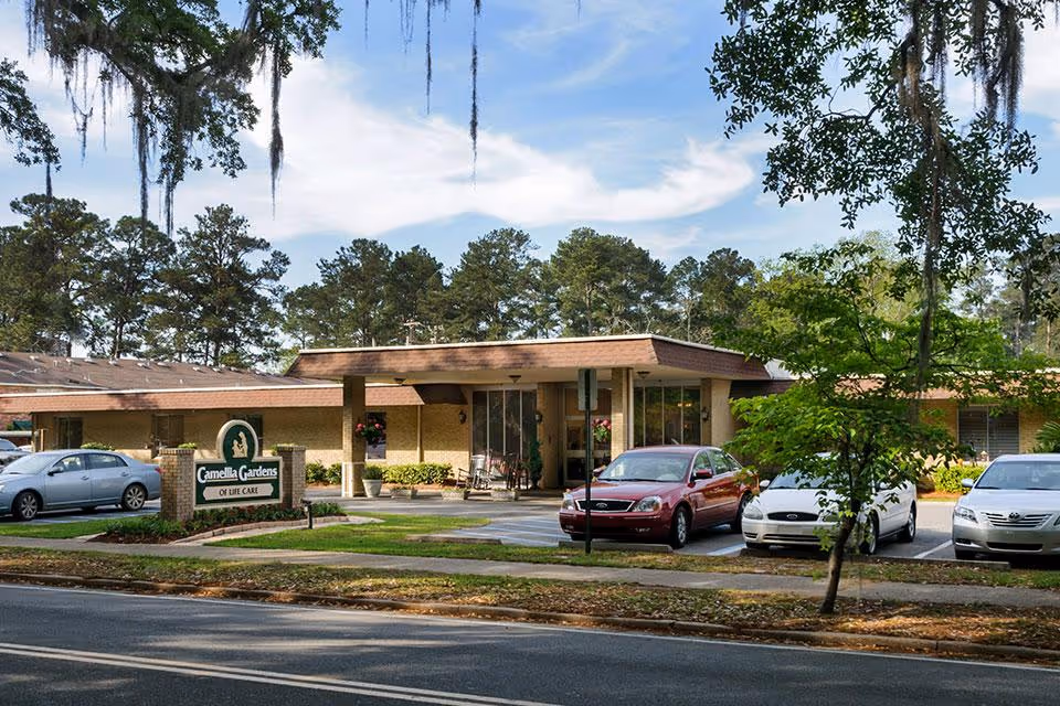 Exterior view of Camellia Gardens of Life Care facility showing a single-story building with a brown roof, several parked cars in front, a sign with the facility name, and trees in the background under a partly cloudy sky.