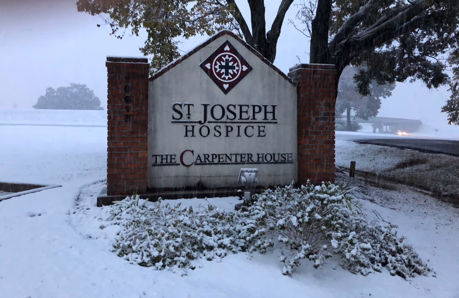 Snow-covered sign reading St. Joseph Hospice stands between brick pillars in a winter landscape.