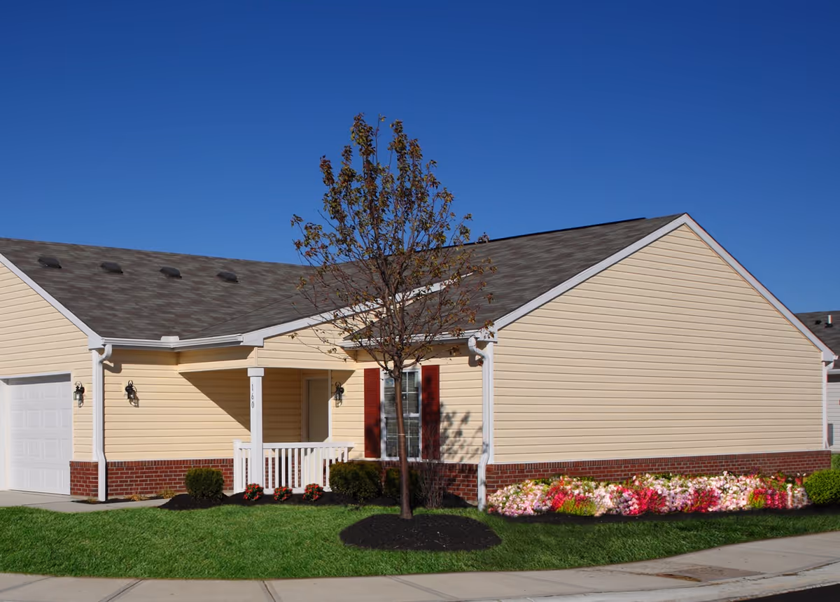 Single-story beige vinyl-sided house with an attached garage, small front porch, tree and colorful flower beds under a clear blue sky.