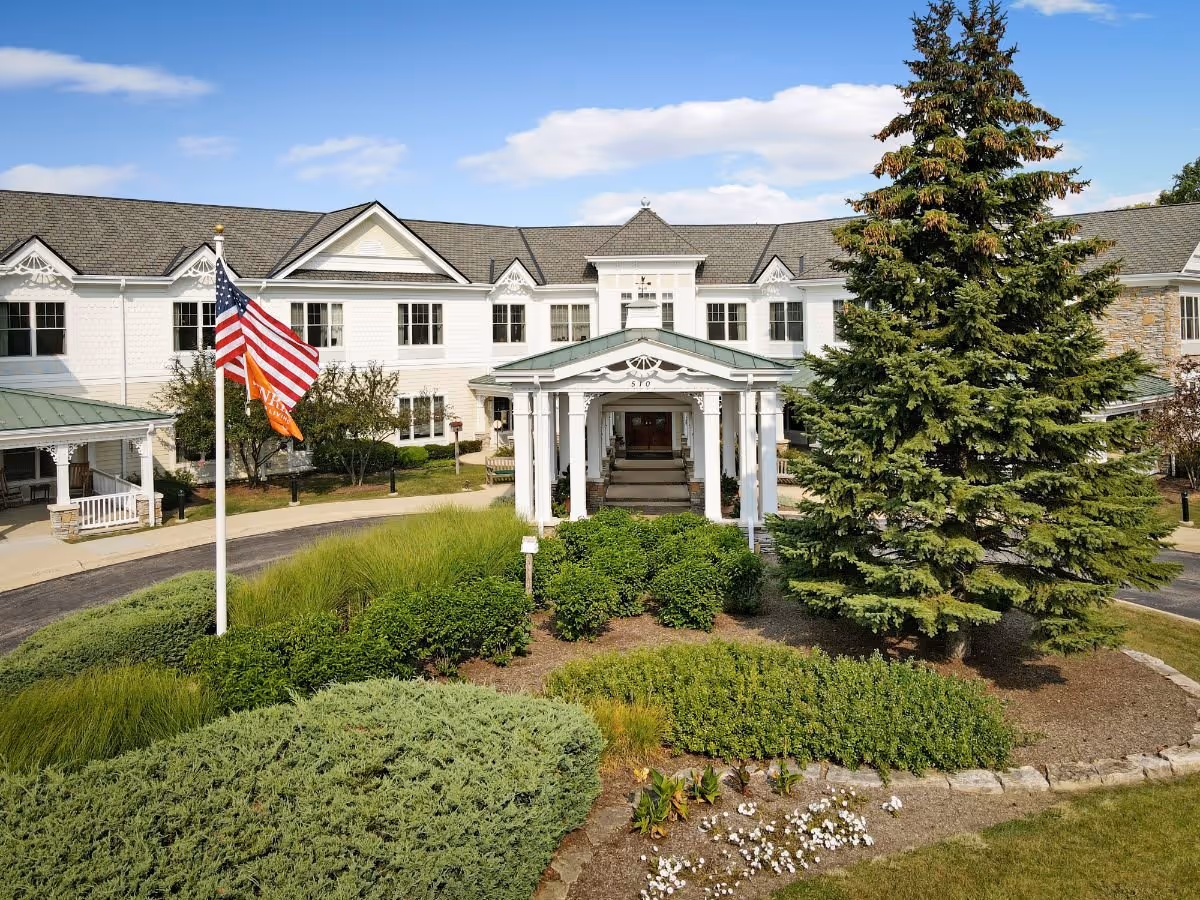 Front entrance of a white two-story senior living building with a porte-cochere, American flag, and landscaped grounds.