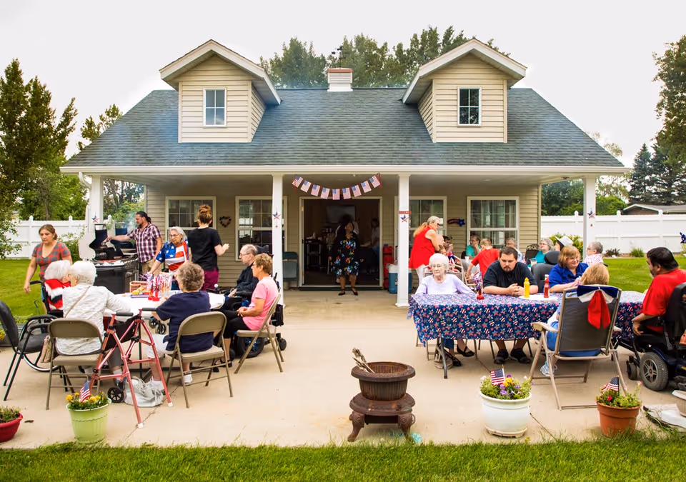 A group of elderly people and caregivers gathered outside a beige house with a covered porch, sitting at tables covered with patriotic-themed tablecloths. Some people are seated in wheelchairs and others in chairs, enjoying a social event with decorations including small American flags and a banner hanging from the porch roof. The setting is a grassy yard with potted plants and a fire pit in the foreground.