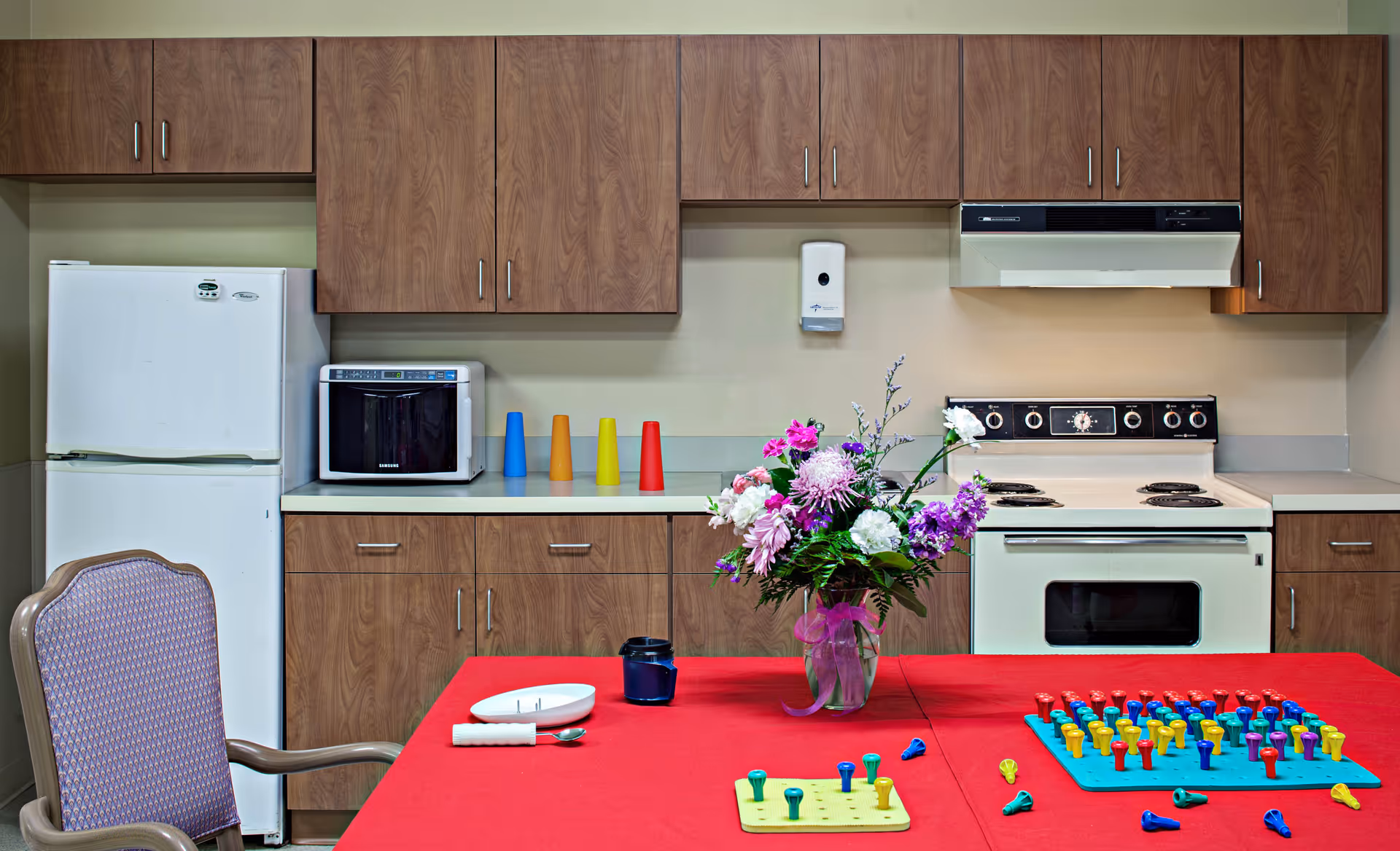 A kitchen area with wooden cabinets, a white refrigerator, microwave, and stove. A table covered with a red tablecloth is in the foreground, featuring a vase of colorful flowers, a blue cup, a white plate with utensils, and pegboards with colorful pegs for activities. A purple cushioned chair is positioned at the table.