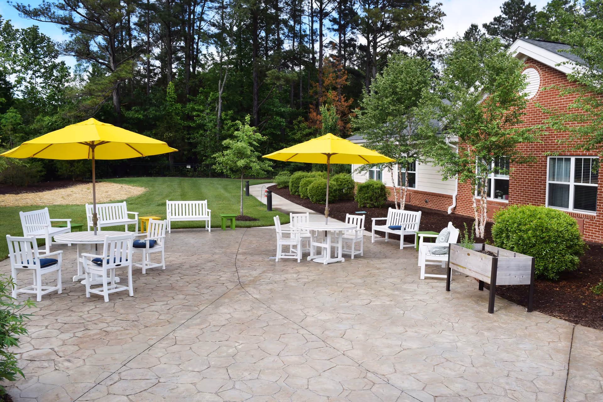 Outdoor patio area with white wooden tables and chairs arranged under two large yellow umbrellas. The patio is paved with stone tiles and surrounded by green grass, bushes, and trees. A red brick building with white trim is visible on the right side of the image.