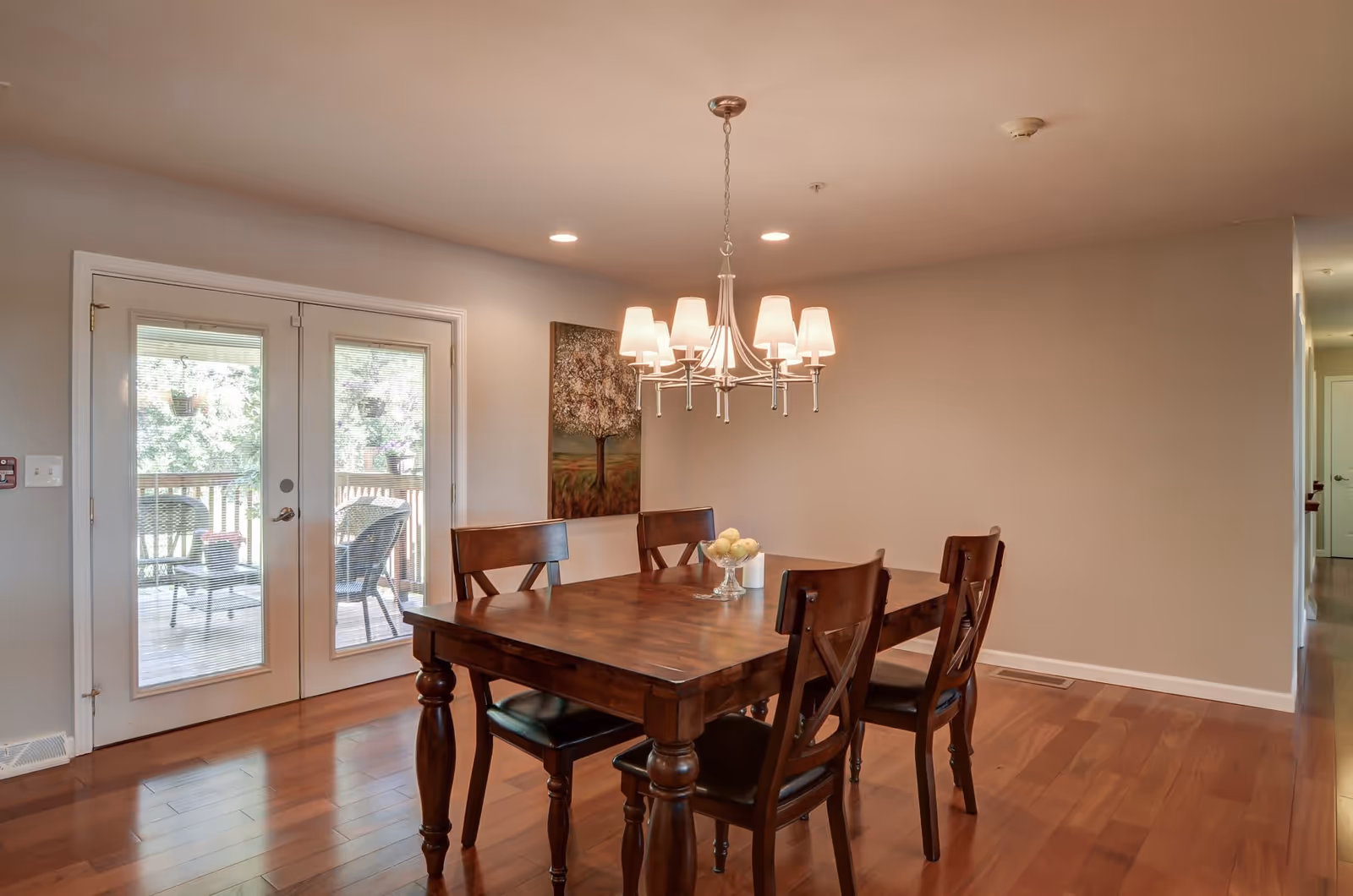 A dining room with a wooden table and four matching chairs on a polished hardwood floor. Above the table hangs a chandelier with multiple lampshades. There is a painting of a tree on the wall and double glass doors leading to an outdoor patio with chairs and potted plants.
