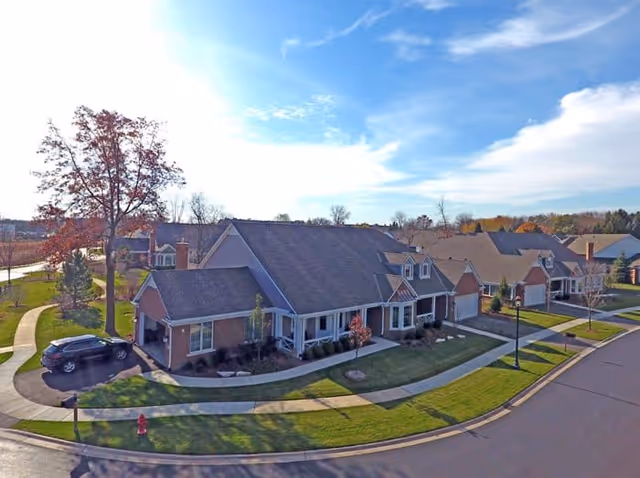 Aerial view of a residential-style senior living facility with multiple connected units featuring pitched roofs, front porches, and garages. The buildings are surrounded by well-maintained lawns, sidewalks, and a few trees under a partly cloudy blue sky.