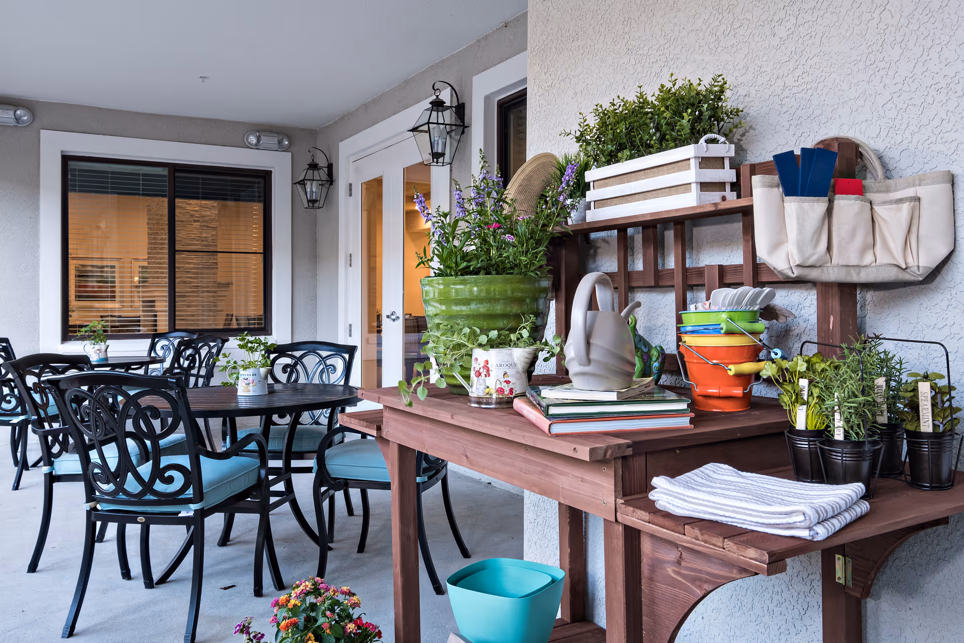 Covered outdoor patio area with black metal tables and chairs with blue cushions. A wooden potting bench holds various gardening supplies including potted plants, gardening tools, watering cans, and books. The patio is adjacent to a building with windows and glass doors.
