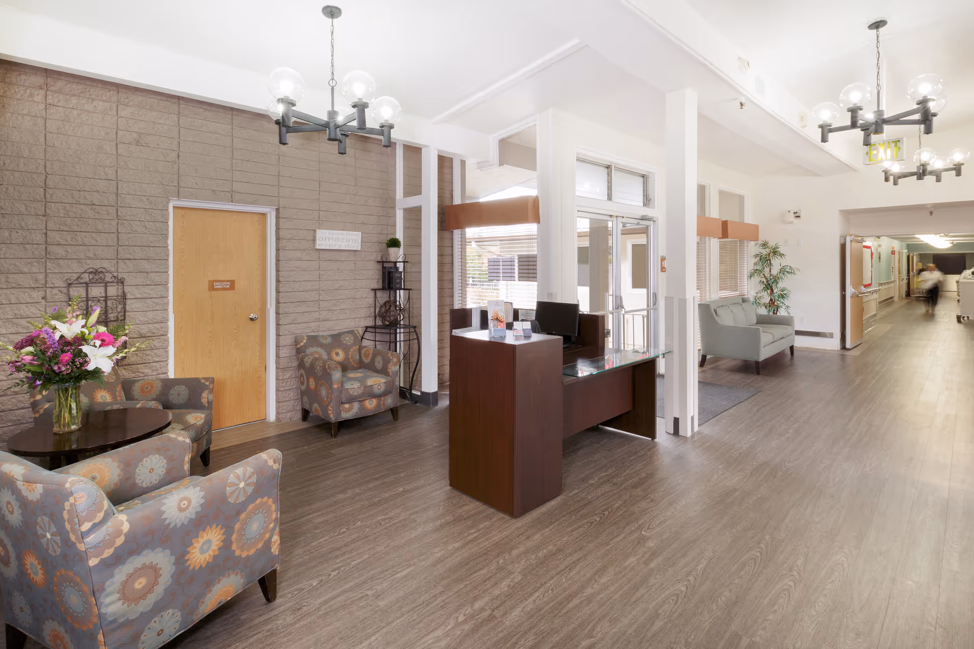 A bright and spacious interior area of a senior living facility with a reception desk, patterned armchairs around a small round table with a flower vase, and a hallway leading to other rooms. The walls are light-colored with a textured accent wall, and there are modern light fixtures on the ceiling.