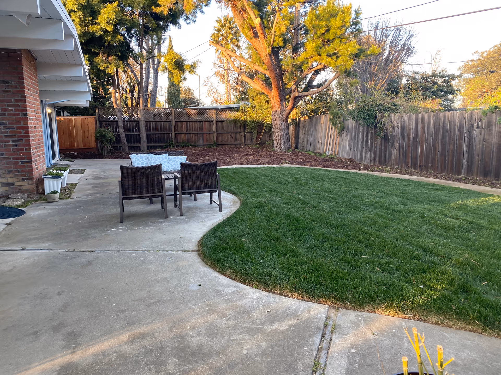 Outdoor patio area with a concrete walkway and a small seating arrangement consisting of two dark wicker chairs and a table with cushions. The patio is adjacent to a brick building and overlooks a well-maintained grassy lawn bordered by a wooden fence and trees in the background.