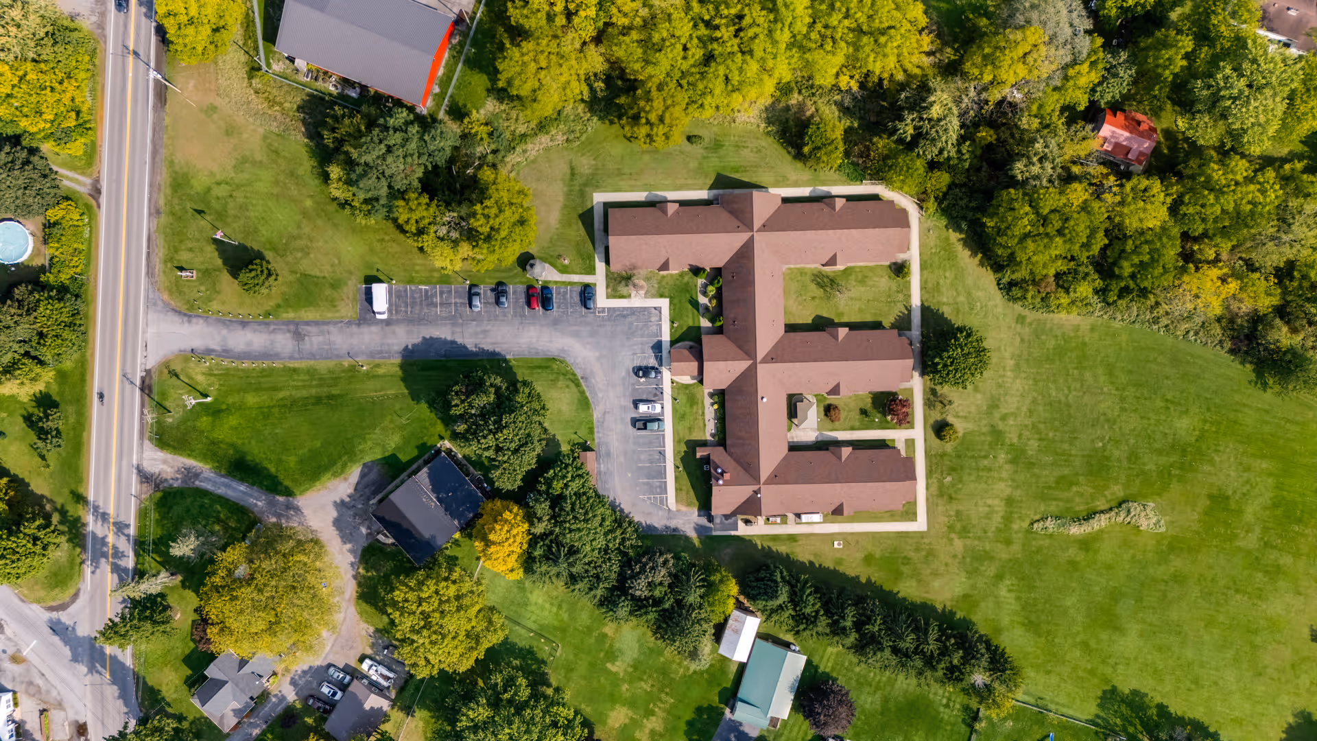 Aerial view of Clinton Crest Manor, showing a large building with a brown roof shaped like the letter E, surrounded by green lawns, trees, and a parking lot with several cars. Nearby roads and additional smaller buildings are also visible.