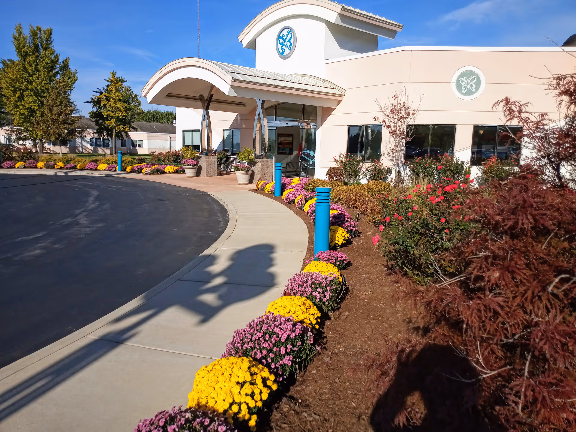 Entrance to a senior living facility with curved canopy, windows, and colorful flower beds.