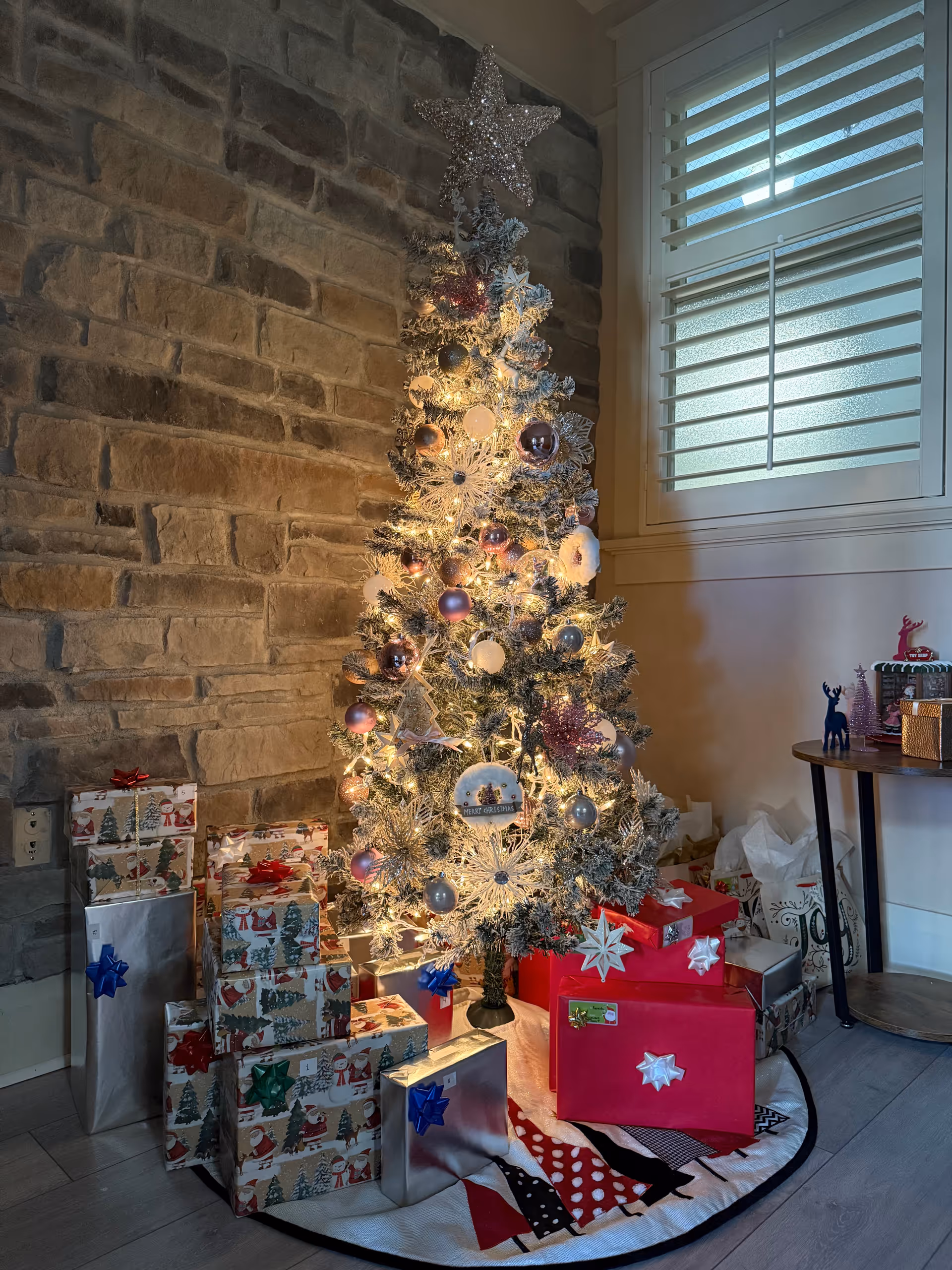 A lit Christmas tree with wrapped presents around it, against a stone wall inside a room.