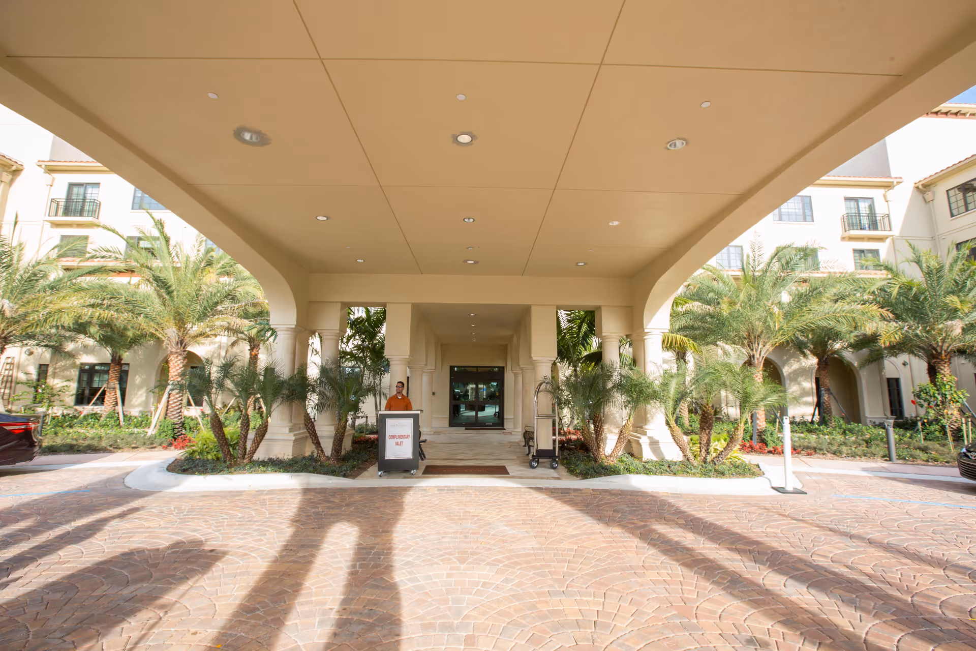 Entrance area of a senior living facility with a covered driveway supported by columns, surrounded by palm trees and landscaping. A person stands near a sign that reads 'COMPLIMENTARY VALET'. The building has multiple floors with balconies and windows.