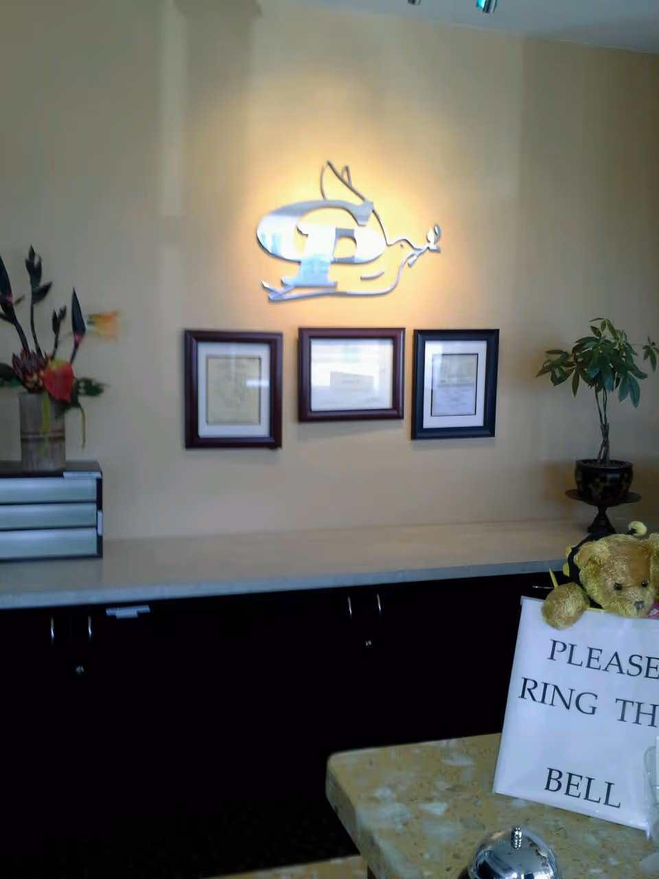 Reception area with logo, framed certificates, plants, a teddy bear, and a bell sign.