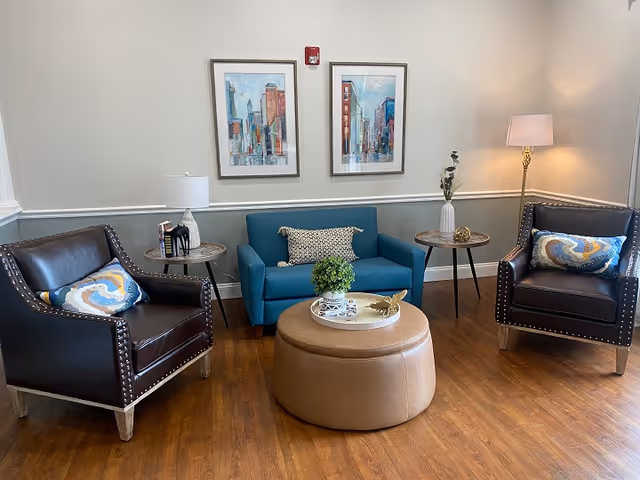 A cozy sitting area with two dark leather armchairs featuring decorative pillows, a small blue loveseat with a patterned pillow, a round tan ottoman in the center, two small round side tables with decorative items, a floor lamp, and two framed cityscape paintings on the wall.