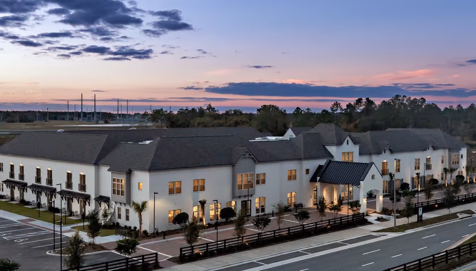 Exterior front view of a two-story senior living facility at dusk with illuminated windows, an entrance canopy, and landscaped grounds.