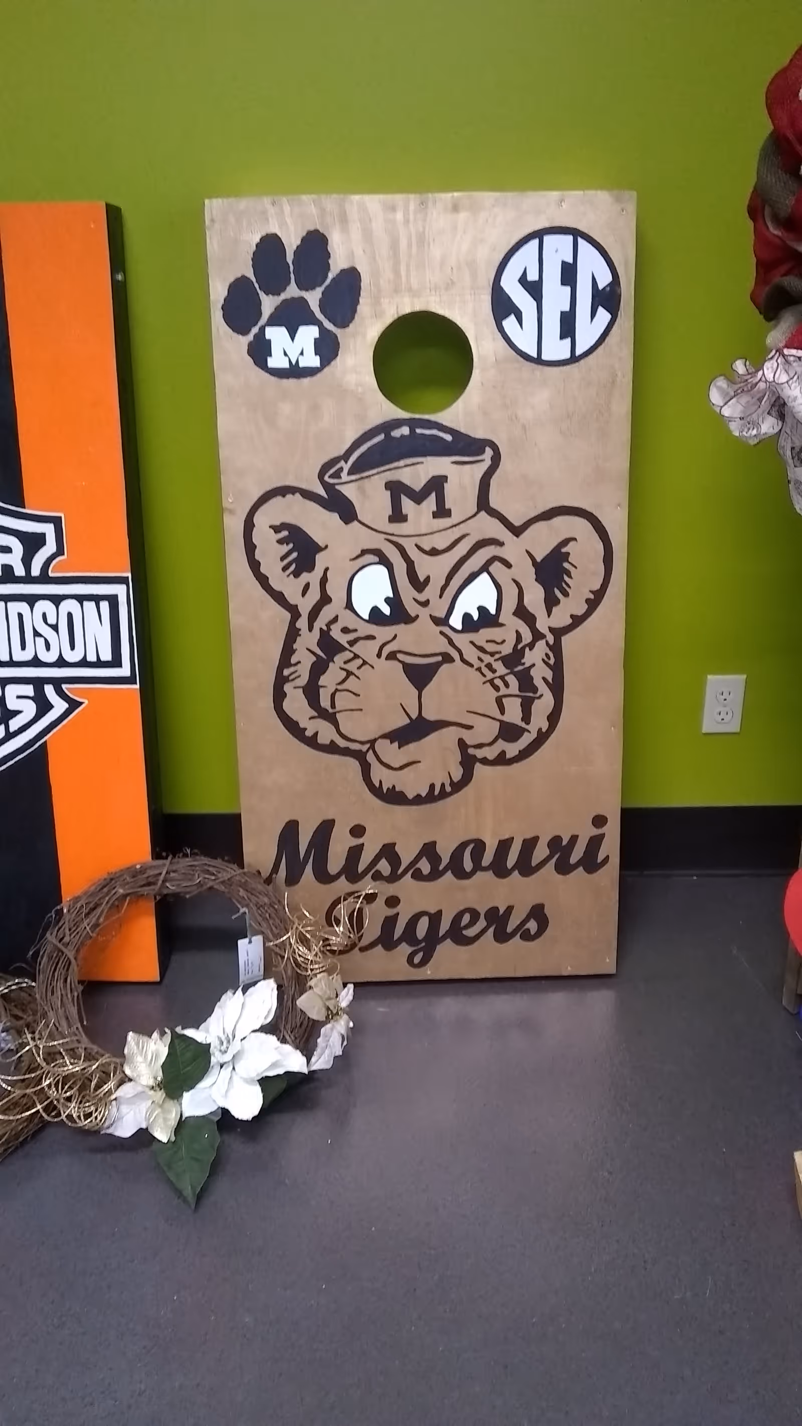 Wooden Missouri Tigers cornhole board leaning against a lime-green wall, with a wreath on floor.