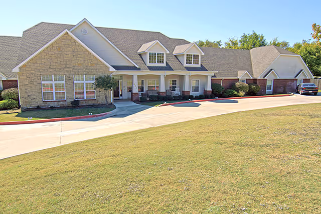 Exterior view of a single-story assisted living facility with a stone and siding facade, a covered porch with chairs, a driveway, and a well-maintained lawn under a clear sky.