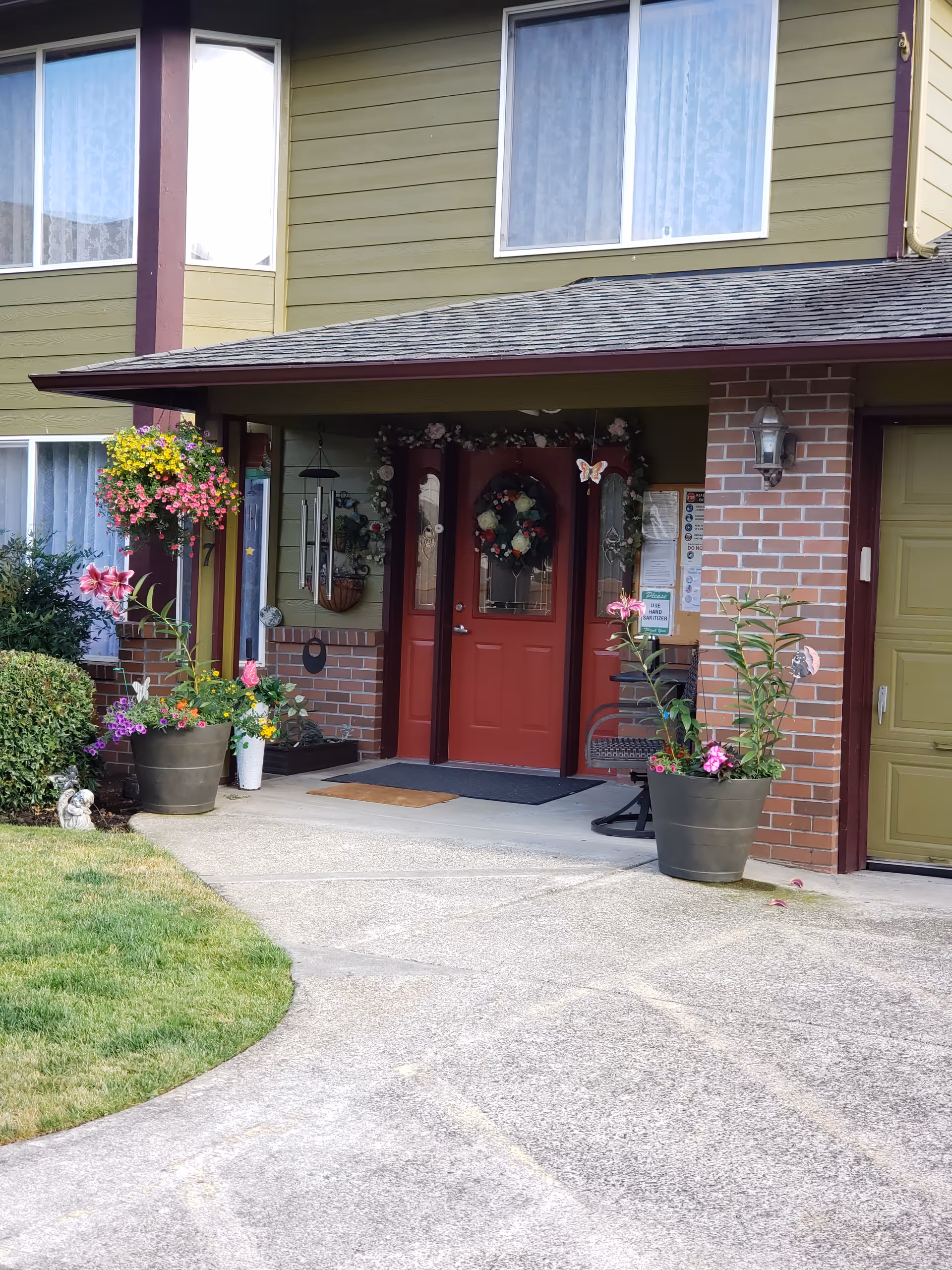 Green siding and brick building entrance with a red door, wreath, and potted flowers.