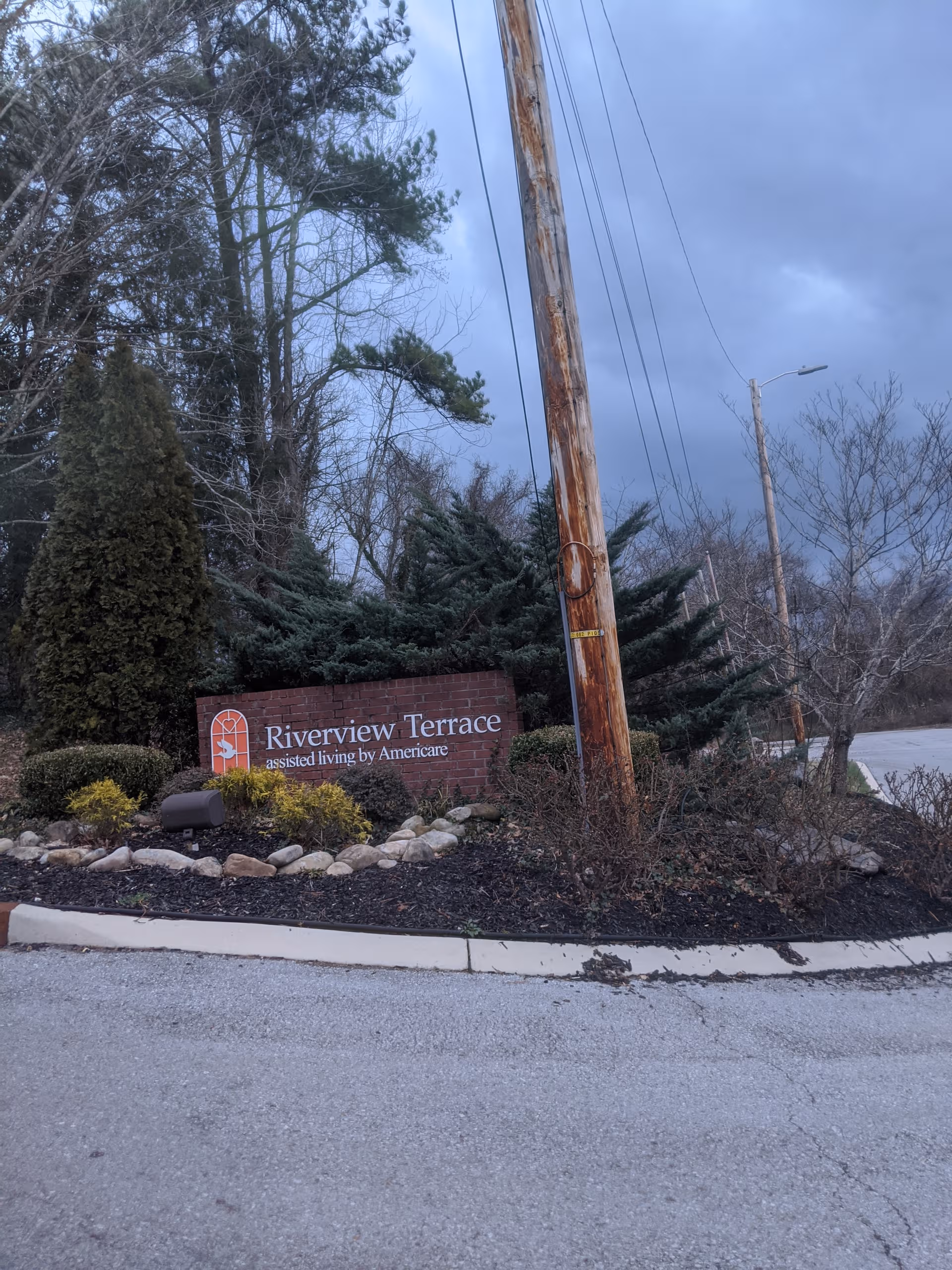 Riverview Terrace entrance sign with landscaping, trees, and a utility pole along the road.